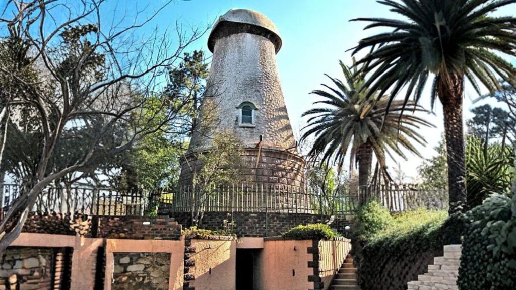 A round, stone windmill tower with a conical roof stands surrounded by palm trees and greenery, enclosed by a metal fence—an inspiring example of homeownership—complete with a brick and stone entrance in the foreground.