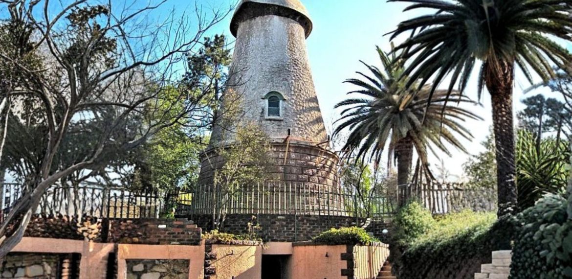 A round, stone windmill tower with a conical roof stands surrounded by palm trees and greenery, enclosed by a metal fence—an inspiring example of homeownership—complete with a brick and stone entrance in the foreground.