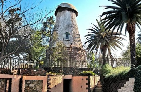 A round, stone windmill tower with a conical roof stands surrounded by palm trees and greenery, enclosed by a metal fence—an inspiring example of homeownership—complete with a brick and stone entrance in the foreground.
