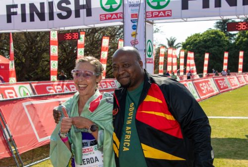 A smiling female runner draped in a flag gives a thumbs up beside a man in a South Africa shirt at the SPAR Womens Challenge race finish line, with timing clocks and banners visible in the background.