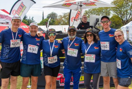 Seven smiling runners in matching blue shirts and medals stand together outdoors after the SPAR Womens Challenge, race bibs on their chests. Colorful event banners and tents are visible in the background.