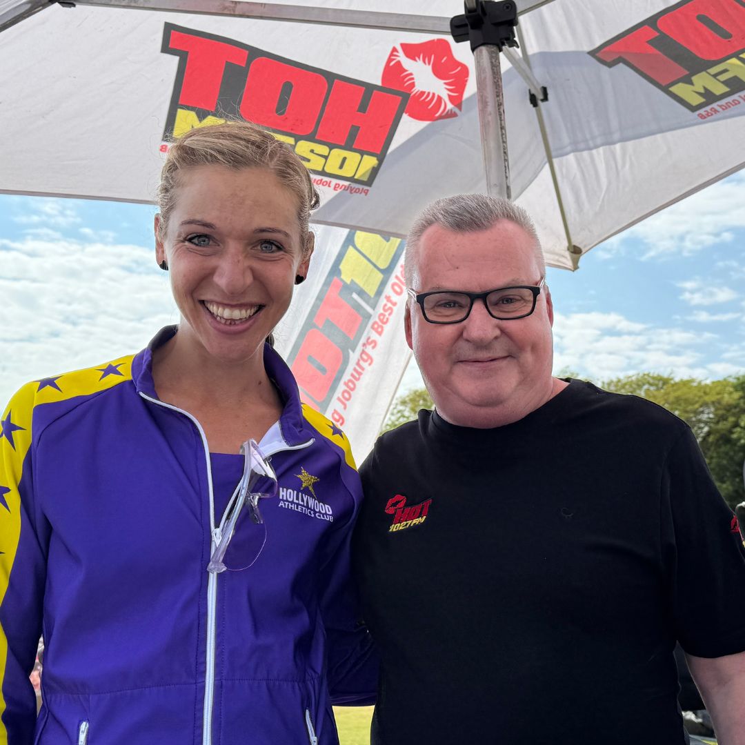 A woman in a purple and yellow jacket stands smiling next to a man in a black shirt, both under a large umbrella with a red and yellow logo, enjoying the sunny day without a hint of brainrot.