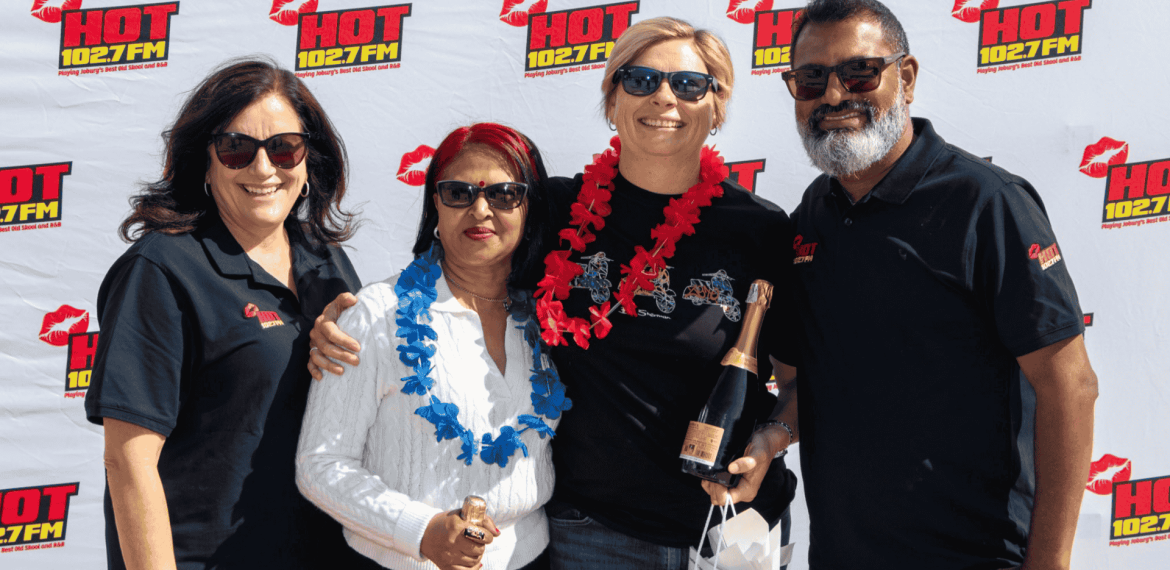 Four people wearing sunglasses and black shirts stand smiling in front of a HOT 102.7 FM backdrop, holding champagne bottles and gift bags. Channeling Jane Goodall’s spirit, two wear flower garlands as they celebrate with happiness and pride.