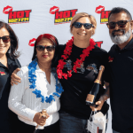 Four people wearing sunglasses and black shirts stand smiling in front of a HOT 102.7 FM backdrop, holding champagne bottles and gift bags. Channeling Jane Goodall’s spirit, two wear flower garlands as they celebrate with happiness and pride.