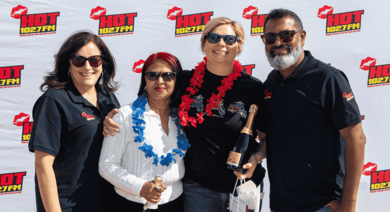 Four people wearing sunglasses and black shirts stand smiling in front of a HOT 102.7 FM backdrop, holding champagne bottles and gift bags. Channeling Jane Goodall’s spirit, two wear flower garlands as they celebrate with happiness and pride.