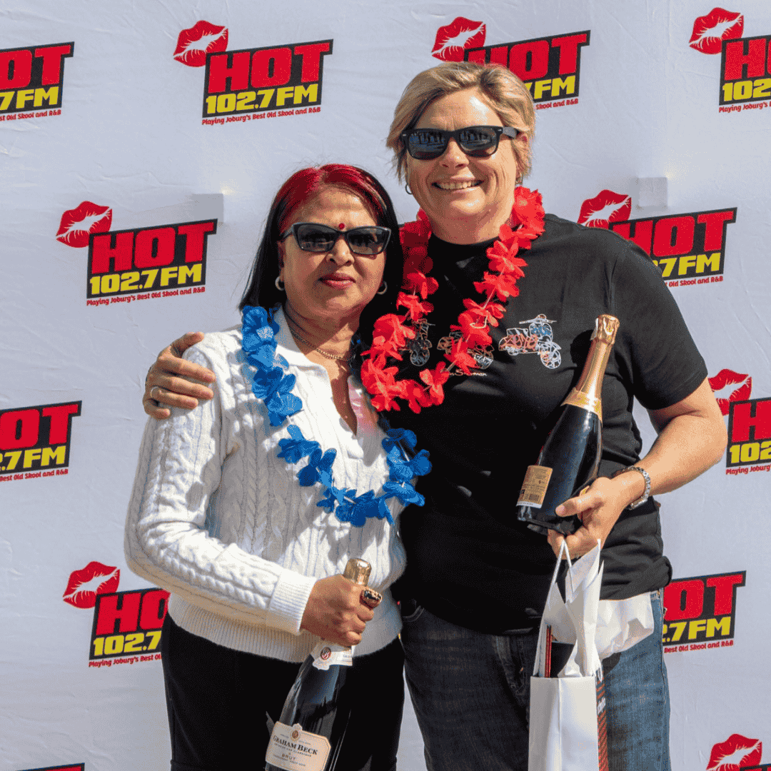 Two women wearing sunglasses and leis, smiling and holding champagne bottles, stand together in front of a HOT 102.7 FM branded backdrop, channeling a celebration-worthy Jane Goodall spirit.