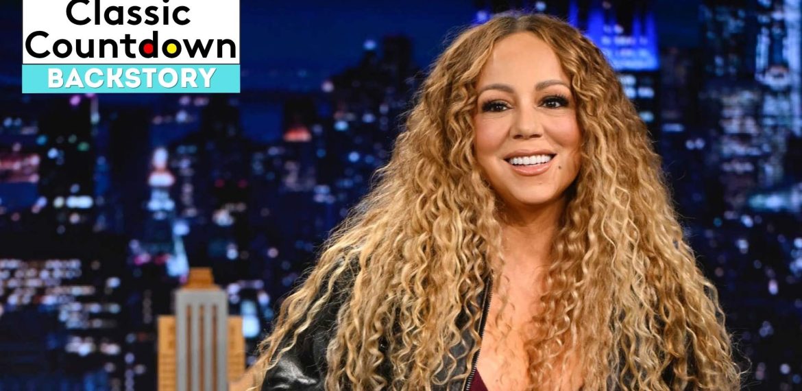Mariah Carey with long curly hair smiles while sitting on a talk show set at night. The text “Classic Countdown Backstory” appears in the top left corner. A cityscape is softly blurred in the background.