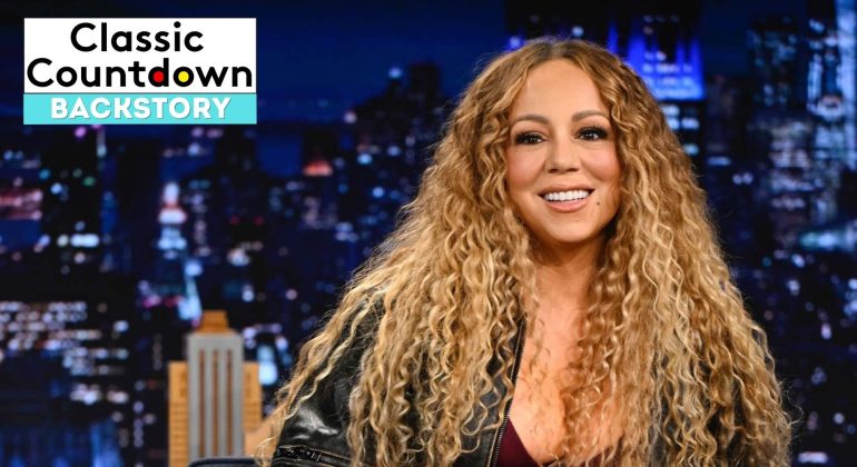 Mariah Carey with long curly hair smiles while sitting on a talk show set at night. The text “Classic Countdown Backstory” appears in the top left corner. A cityscape is softly blurred in the background.
