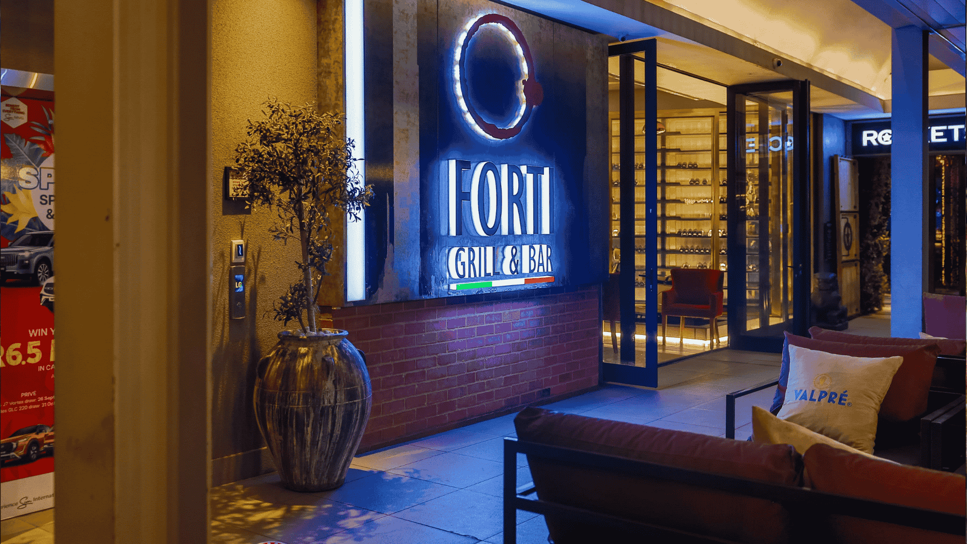 Outdoor view of the illuminated entrance to Fort Grill & Bar, featuring a large sign and modern decor—perfect for celebrating Heritage Day. Seating area with cushions, potted plant, and glass doors reveal a stylish wine rack inside.