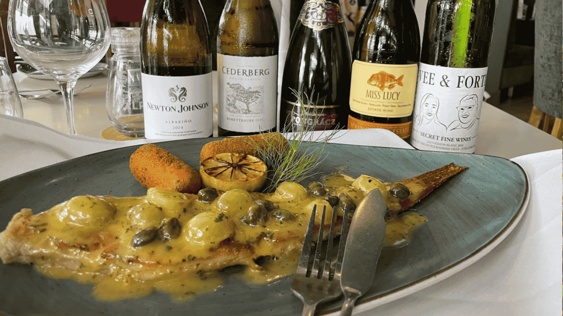 A plate with fish in a creamy sauce, garnished with capers, grilled lemon, and a croquette, is set before five wine bottles and an empty glass on a white tablecloth—perfect for celebrating Heritage Day.
