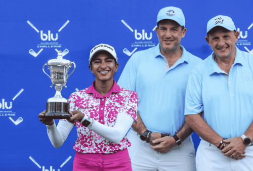 A smiling female golfer in a pink and white outfit holds a trophy, standing before a blue backdrop with Advanced Hair Clinic branding, crossed golf clubs, and the word blu, joined by two men in light blue shirts and hats.
