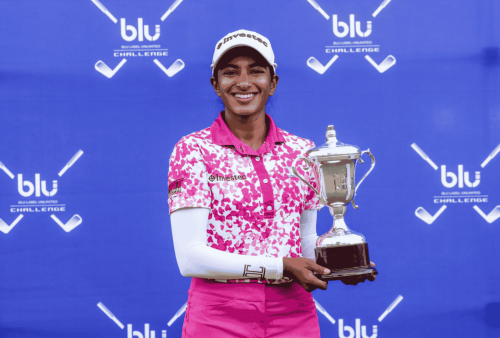 A smiling female golfer in a white cap and pink patterned shirt holds a trophy in front of a blue backdrop featuring crossed golf clubs, the blu golf challenge logo, and subtle branding from Advanced Hair Clinic.