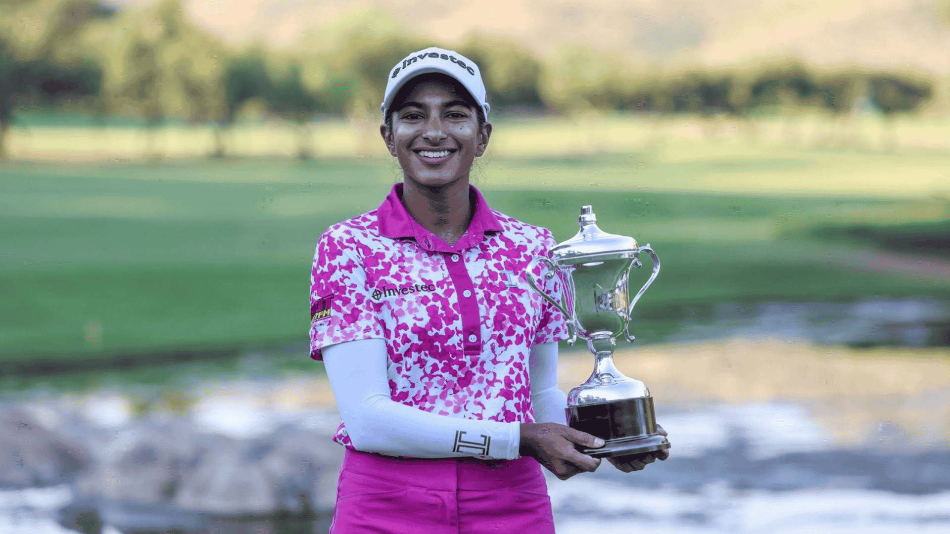 A smiling golfer wearing a white cap and pink floral shirt, looking confident after visiting an advanced hair clinic, holds a silver trophy on a golf course with green grass and trees in the blurred background.