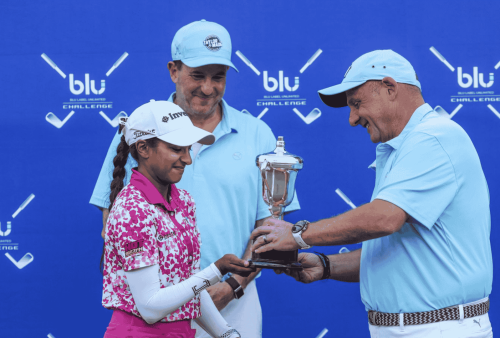 A female golfer in a pink and white outfit receives a trophy from a man in a blue shirt and cap, with another man in blue standing behind them. The blue backdrop features blu challenge and Advanced Hair Clinic logos.