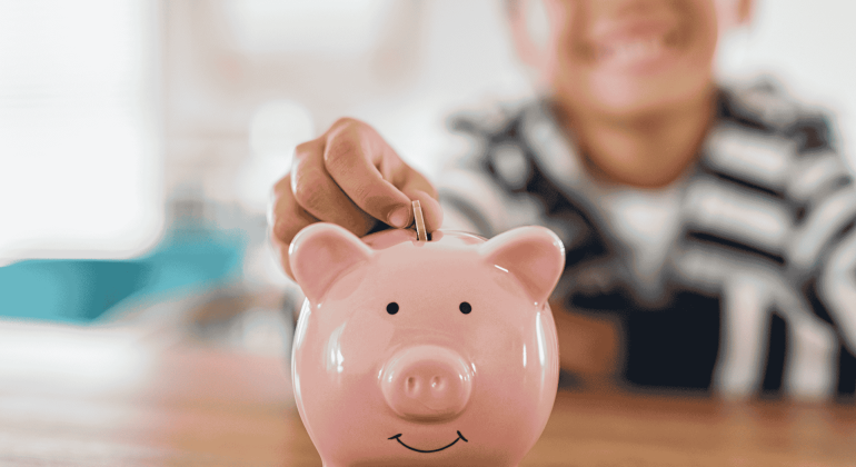 A child smiles while placing a coin into a pink piggy bank on a wooden table, showing that saving money is far from brainrot. The background is softly blurred.
