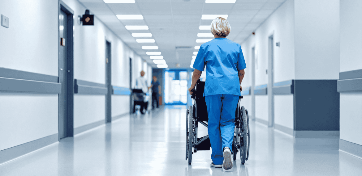 A healthcare worker in blue scrubs pushes an empty wheelchair down a bright, clean hospital corridor, their brisk pace breaking through the brainrot of a long shift. Other people are visible in the distance at the end of the hallway.