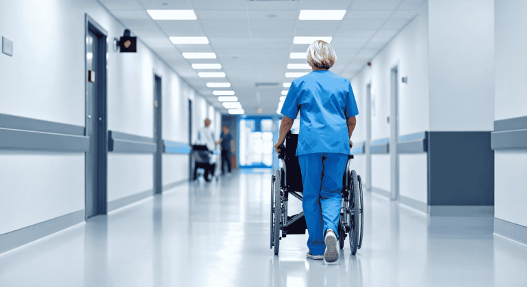 A healthcare worker in blue scrubs pushes an empty wheelchair down a bright, clean hospital corridor, their brisk pace breaking through the brainrot of a long shift. Other people are visible in the distance at the end of the hallway.