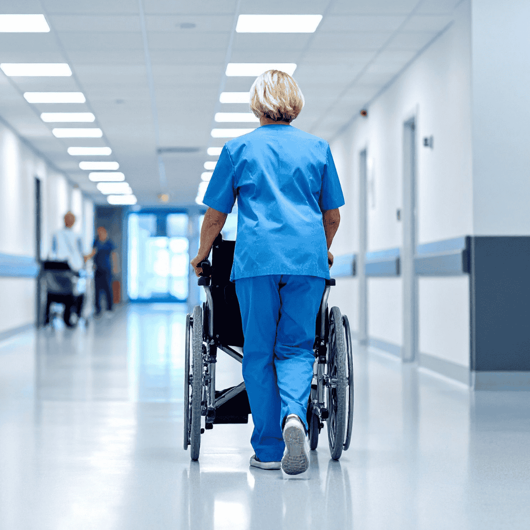 A healthcare worker in blue scrubs walks down a hospital hallway, pushing an empty wheelchair. The brightly lit, clean hall is free from chaos or brainrot, with doors and lights neatly lining the walls.