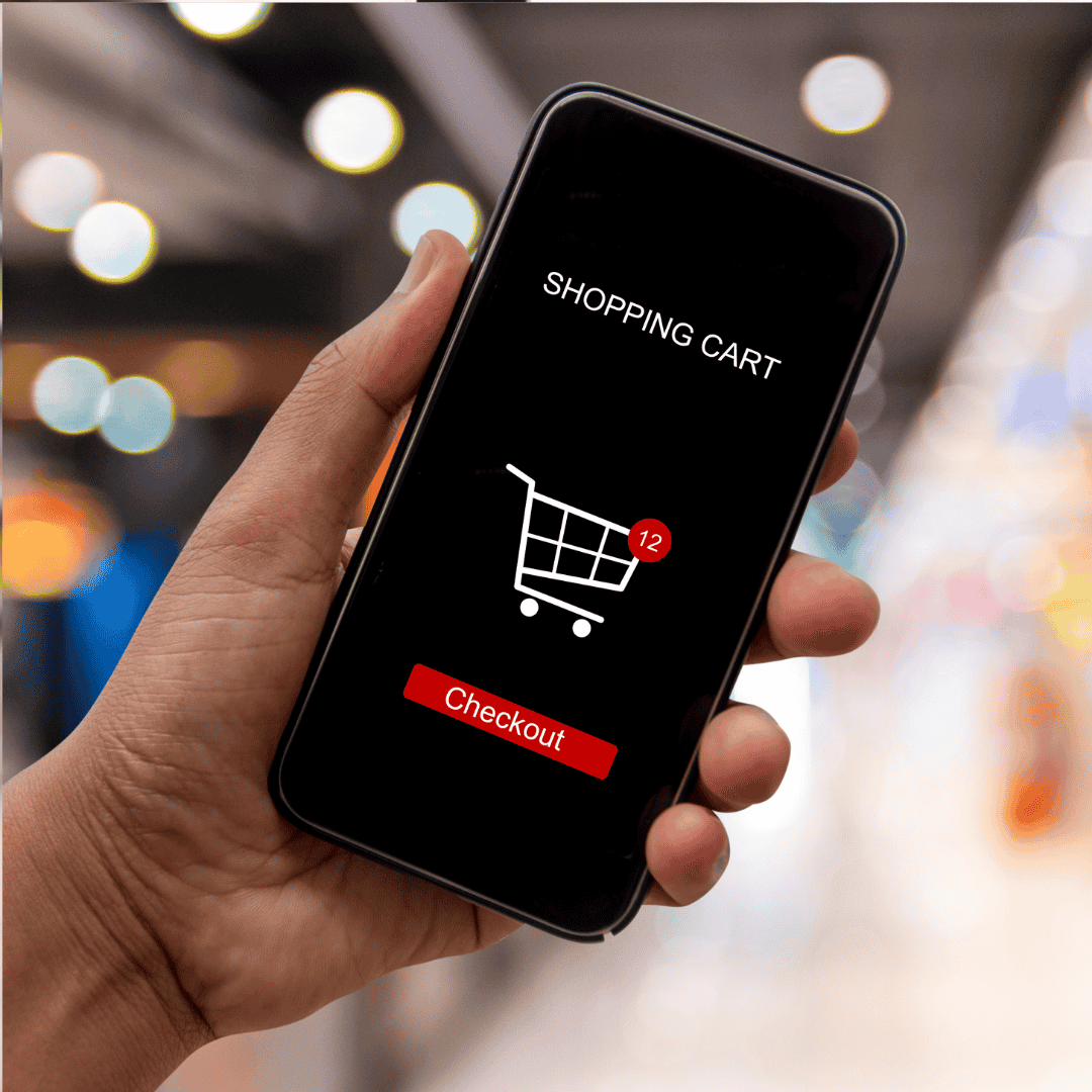 A hand holds a smartphone displaying a shopping cart icon with 12 items and a red Checkout button, suggesting the convenience of online shopping—a modern perk of homeownership. The background is blurred with colorful lights.