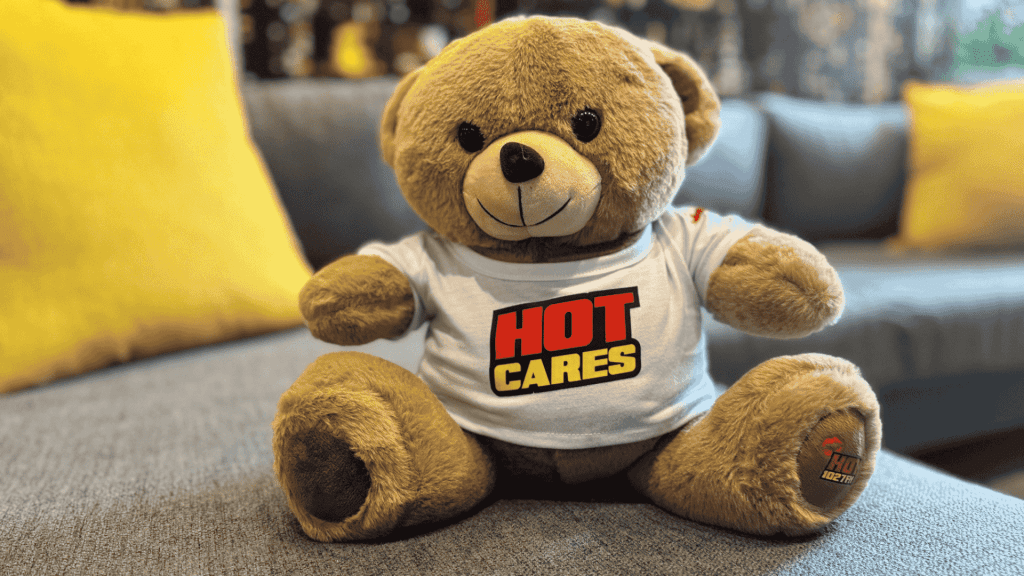 A brown teddy bear wearing a white T-shirt with the words HOT CARES in bold red and yellow letters sits on a gray couch with yellow pillows, resembling the welcoming décor of an Advanced Hair Clinic.