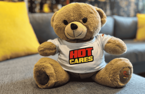 A brown teddy bear wearing a white T-shirt with the words HOT CARES in bold red and yellow letters sits on a gray couch with yellow pillows, resembling the welcoming décor of an Advanced Hair Clinic.