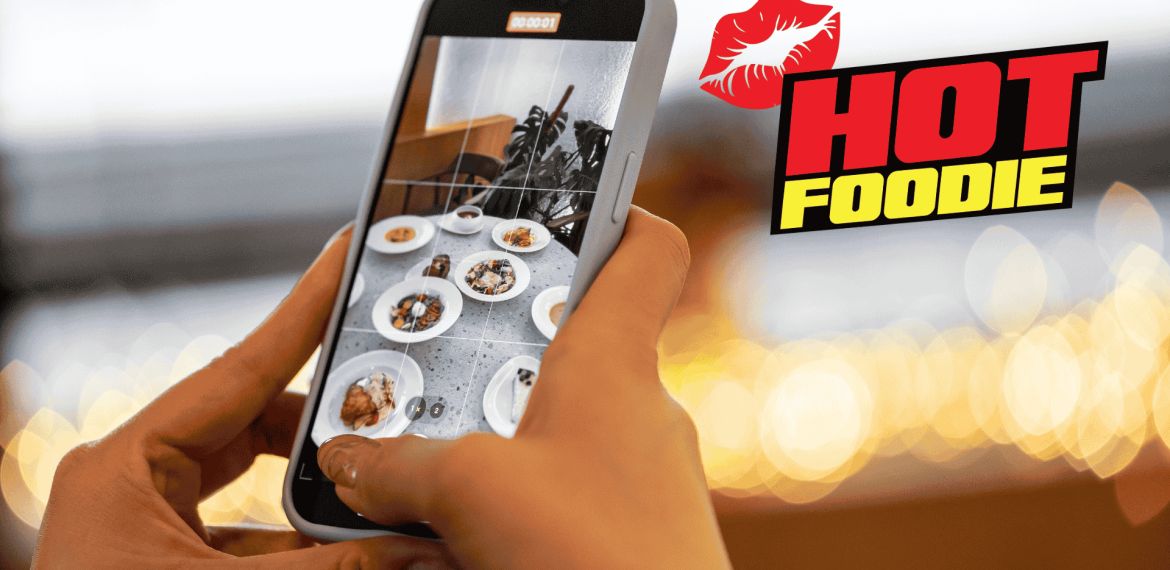 A person takes a photo of various plated dishes with a smartphone. The background is blurred lights, and the words HOT FOODIE with a red lips graphic appear on the right side, celebrating heritage day flavors.