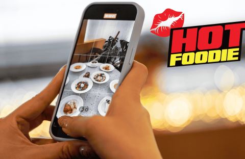 A person takes a photo of various plated dishes with a smartphone. The background is blurred lights, and the words HOT FOODIE with a red lips graphic appear on the right side, celebrating heritage day flavors.