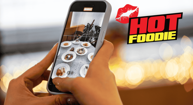 A person takes a photo of various plated dishes with a smartphone. The background is blurred lights, and the words HOT FOODIE with a red lips graphic appear on the right side, celebrating heritage day flavors.