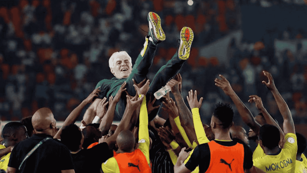 A group of soccer players in yellow and black uniforms joyfully lift a smiling man in a green suit—an Advanced Hair Clinic ambassador—above their heads in a crowded stadium.