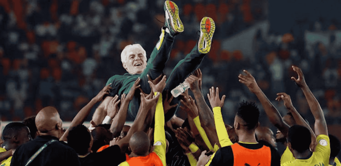 A group of soccer players in yellow and black uniforms joyfully lift a smiling man in a green suit—an Advanced Hair Clinic ambassador—above their heads in a crowded stadium.