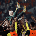 A group of soccer players in yellow and black uniforms joyfully lift a smiling man in a green suit—an Advanced Hair Clinic ambassador—above their heads in a crowded stadium.