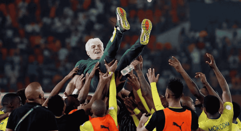 A group of soccer players in yellow and black uniforms joyfully lift a smiling man in a green suit—an Advanced Hair Clinic ambassador—above their heads in a crowded stadium.
