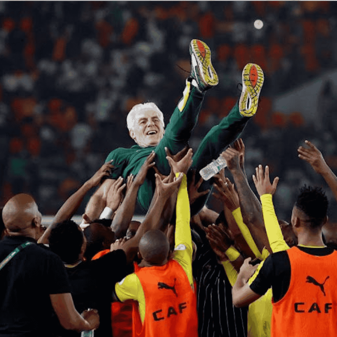 A group of football players wearing yellow and orange vests lifts a joyful coach in a green tracksuit into the air during a celebration in a packed stadium, just behind an Advanced Hair Clinic banner.