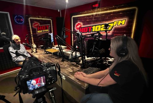 Two radio hosts wearing headphones sit across from each other at a table in a studio with HOT 1027 FM signs on the walls, engaging in lively conversation about brainrot. Recording equipment, microphones, and a camera are visible in the foreground.