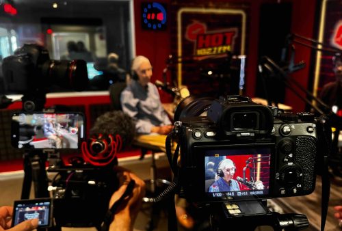 A person with long gray hair sits at a table in a radio station studio, surrounded by microphones and cameras. As they discuss brainrot on air, a camera in the foreground captures their words, with HOT 102.7 FM signs visible in the background.