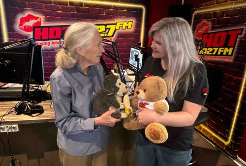 Two women stand in a radio studio, smiling at each other while holding stuffed animals—a monkey and a bear. With microphones and HOT 102.7 FM signs behind them, their playful energy brings some light relief from everyday brainrot.