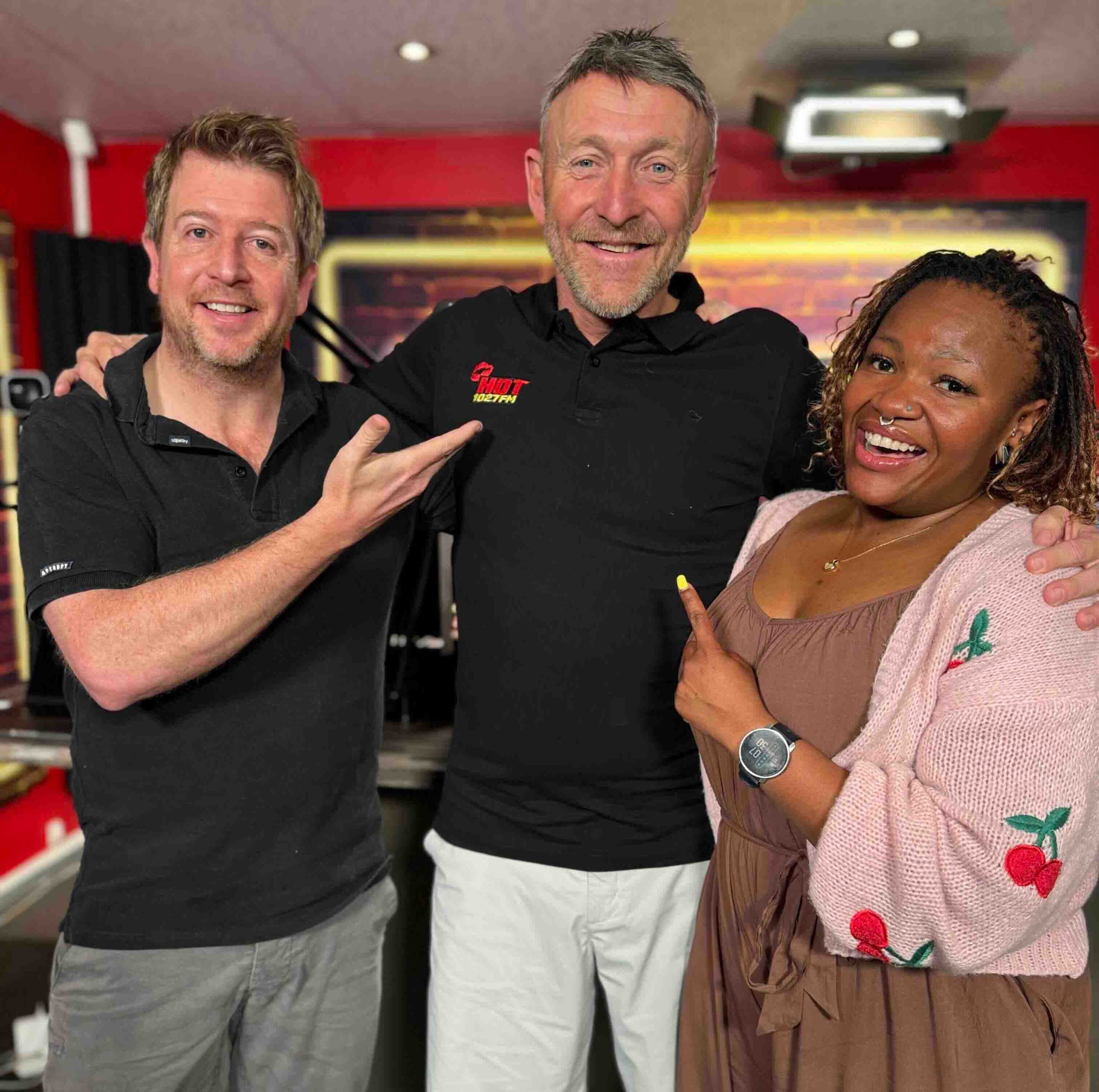 Three people stand smiling in a studio. The two on either side are pointing at the man in the center, who is wearing a black polo shirt with red text. The blurred red and black backdrop hints at their shared passion for homeownership.