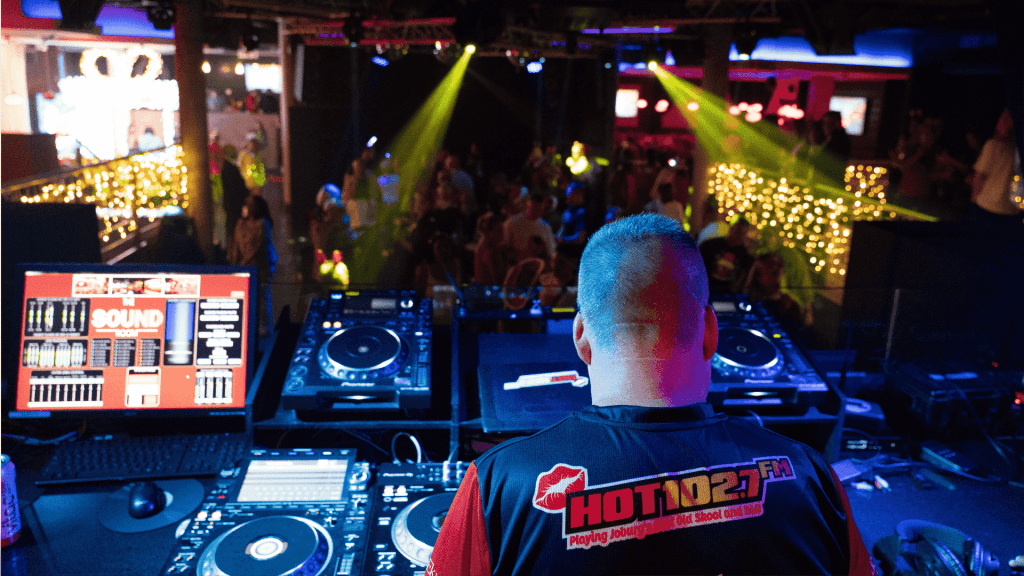 A DJ wearing a HOT 102.7 FM shirt stands at a booth with turntables and computer screens, performing for a lively Jingle Jam crowd in a club lit with colorful lights and string decorations.