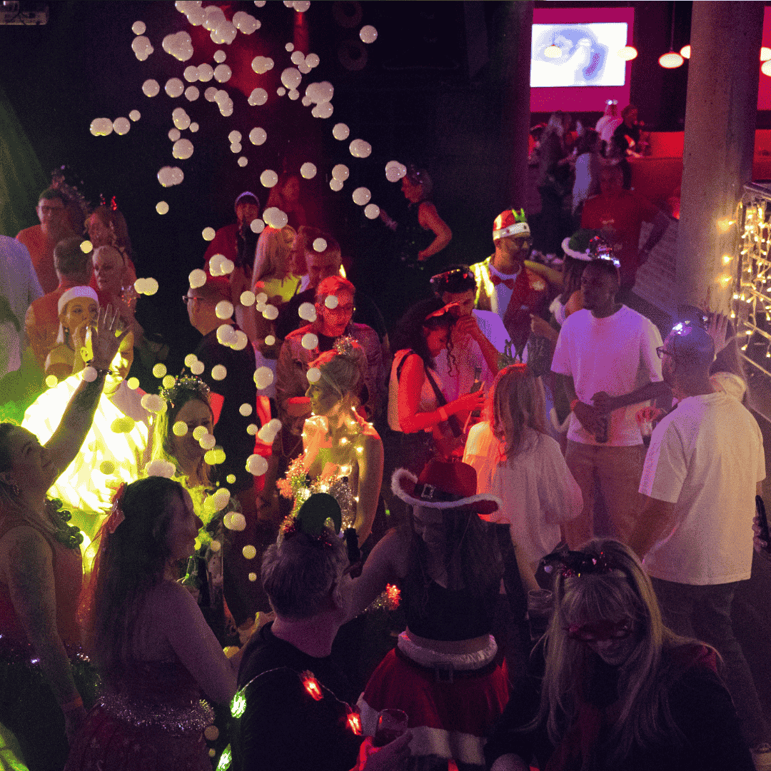 A large group of people in festive attire, some wearing Santa hats and costumes, gather at a lively indoor Jingle Jam party with dim lighting and bright decorative lights hanging above.
