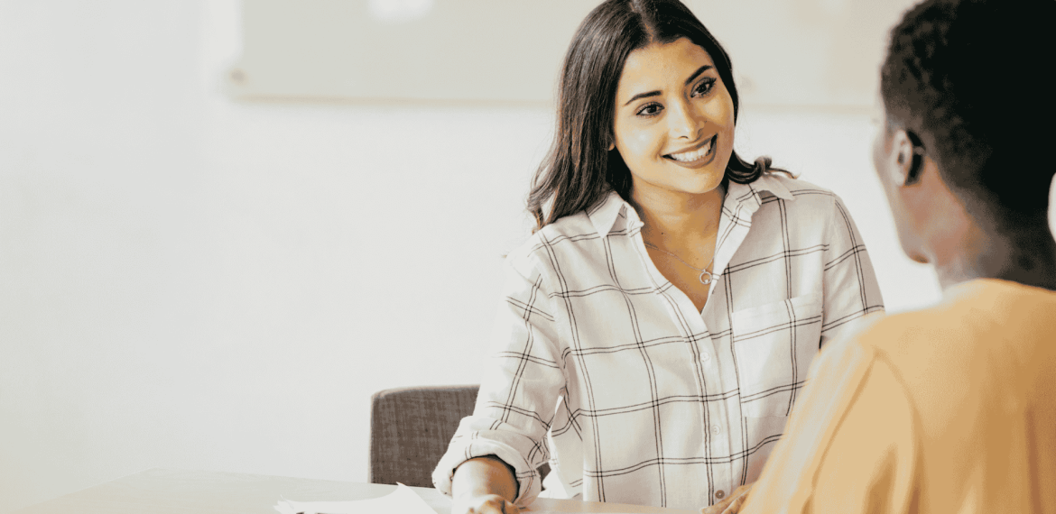 A woman in a checkered shirt smiles while talking to another person across a table, a clipboard and pen in front of her, sharing a lighthearted moment that chases away any hint of brainrot from their busy day.