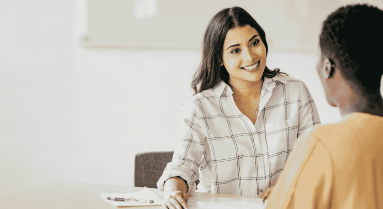 A woman in a checkered shirt smiles while talking to another person across a table, a clipboard and pen in front of her, sharing a lighthearted moment that chases away any hint of brainrot from their busy day.