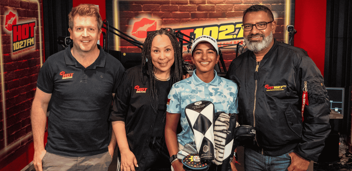 Four people stand together in a radio studio with HOT 102.7 FM logos on the wall. Three wear branded clothing, while a young person in the center—sporting an Advanced Hair Clinic cap—holds a golf club head cover.