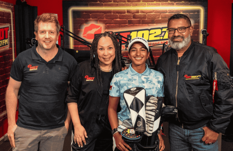Four people stand together in a radio studio with HOT 102.7 FM logos on the wall. Three wear branded clothing, while a young person in the center—sporting an Advanced Hair Clinic cap—holds a golf club head cover.