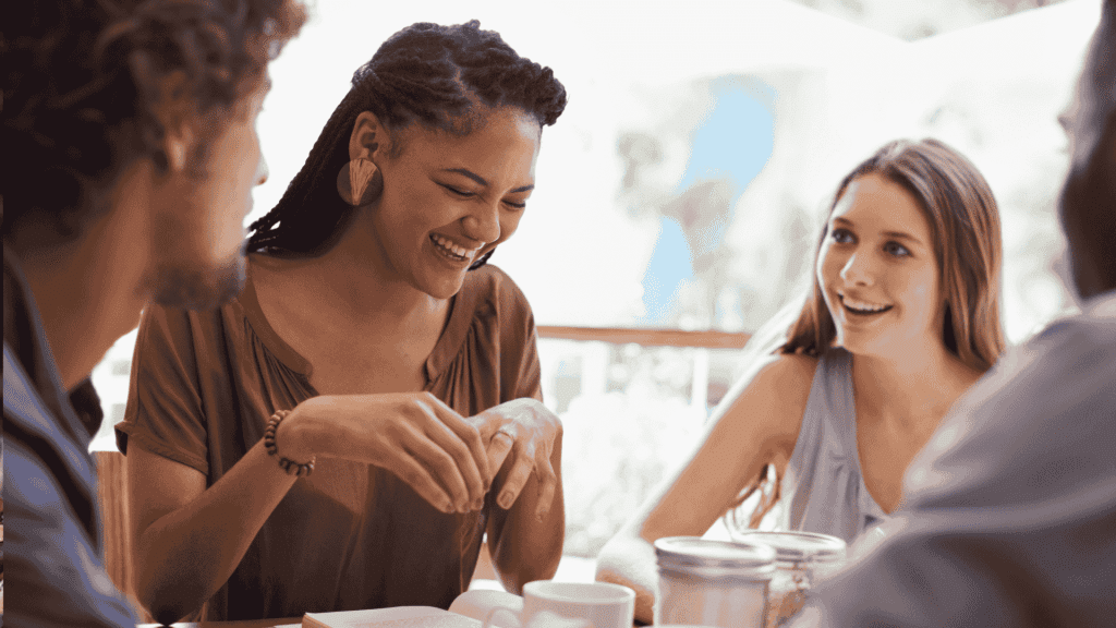 Four people sit together at a table, smiling and talking. One woman in the center, who recently visited an advanced hair clinic, is laughing, while the others listen and engage in conversation in a bright, casual setting.