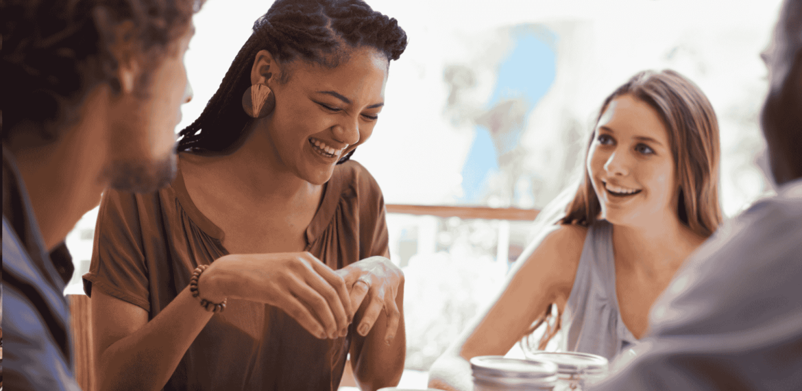 Four people sit together at a table, smiling and talking. One woman in the center, who recently visited an advanced hair clinic, is laughing, while the others listen and engage in conversation in a bright, casual setting.