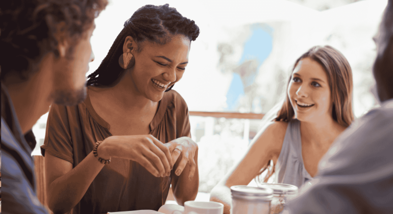 Four people sit together at a table, smiling and talking. One woman in the center, who recently visited an advanced hair clinic, is laughing, while the others listen and engage in conversation in a bright, casual setting.