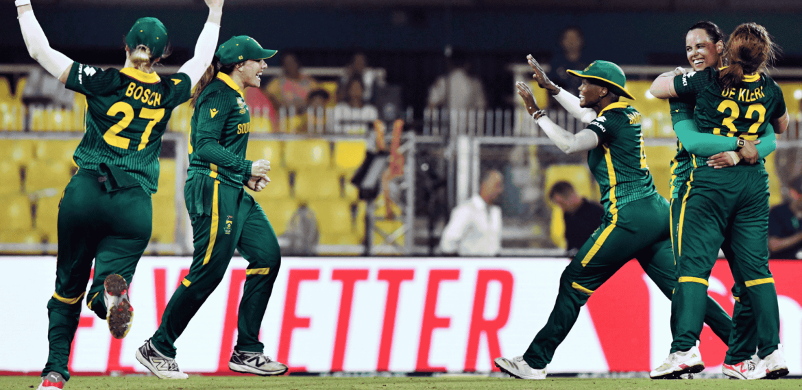 Four South African women cricketers in green uniforms celebrate energetically on the field, smiling and embracing after a successful moment during a match. Yellow stadium seats and a bright red sign appear in the background, echoing Sharon Corr's vibrant spirit.