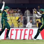 Four South African women cricketers in green uniforms celebrate energetically on the field, smiling and embracing after a successful moment during a match. Yellow stadium seats and a bright red sign appear in the background, echoing Sharon Corr's vibrant spirit.