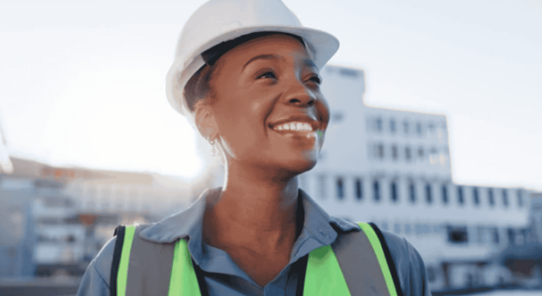 A smiling person wearing a white safety helmet, neon green reflective vest, and blue shirt stands outdoors with buildings in the background, showing no signs of brainrot as they enjoy a clear, focused day at work.