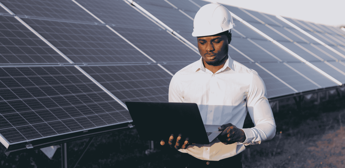A man wearing a white hard hat and shirt stands outdoors in front of solar panels, holding and working on a laptop to ensure efficient service delivery.
